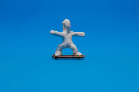 A white plasticine doll practicing skateboarding on a blue background. The doll is balancing on the skateboard made of corrugated cardboard.の写真素材