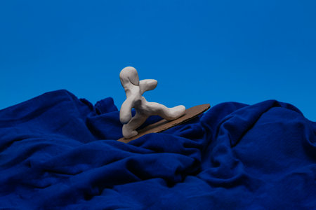 A white plasticine doll surfing on a blue background. The doll is balancing on a corrugated cardboard surf board on waves made of blue fabric.の写真素材