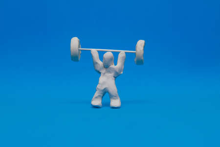 A white plasticine dummy practicing weightlifting on a blue background. The dummy is holding weights above his body by grabbing them from a barbell.の写真素材