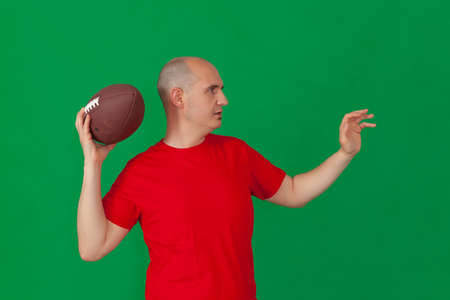 A bald caucasian man dressed in a red t-shirt holding an american football ball positioned as if to throw a forward pass. The background is green.の写真素材