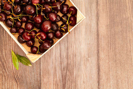 On some wooden boards, in the upper left corner there is a wooden box full of dark red or maroon cherries, next to it there are some green leaves. To the right there is space to fill.の写真素材