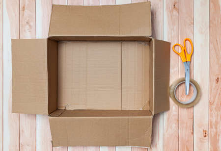 On some wooden boards is an empty cardboard box, ready to be filled or to prepare a package for shipping. There is also a pair of scissors with an orange handle and a roll of transparent sealing tape.の写真素材