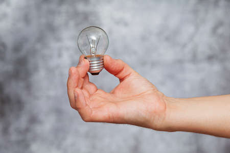 A man's right hand holding an extinguished tungsten filament light bulb on a textured gray background.の写真素材