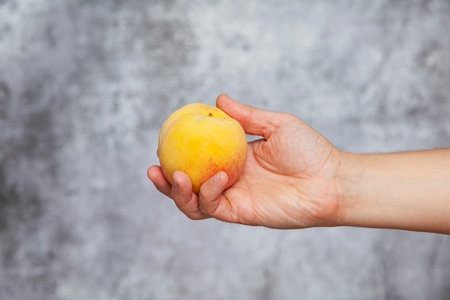 A man's right hand holding a yellow peach on a textured gray background.の写真素材
