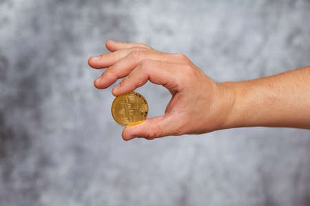 A man's right hand holding a golden Bitcoin coin on a textured gray background.の写真素材