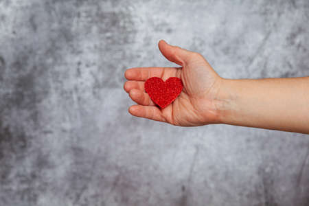 A man's right hand holding a red glitter heart shape on a textured gray background.の写真素材