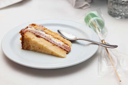 On a table with a white tablecloth is a plate with a slice of cake and a metal spoon. Next to the...