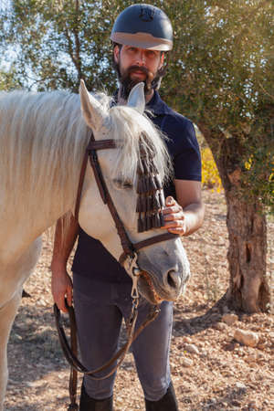 A bearded Caucasian man in equestrian clothing and helmet is standing still stroking the head of his white horse in a field with olive trees.の写真素材
