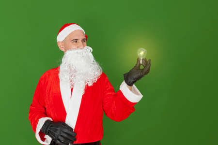 A man dressed as Santa Claus is holding a light bulb with one hand that is lit and emitting light. The background is green.の写真素材