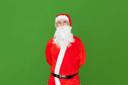 A Caucasian man dressed as Santa Claus looking straight ahead while hiding his hands behind his back. The background is green.の写真素材