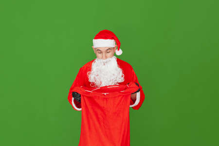 A Caucasian man dressed as Santa Claus is holding a red cloth sack open while looking inside with a surprised expression. The background is green.の写真素材