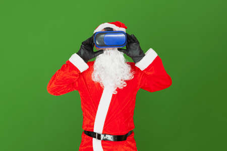 A Caucasian man dressed as Santa Claus is adjusting blue virtual reality goggles. The background is green.の写真素材
