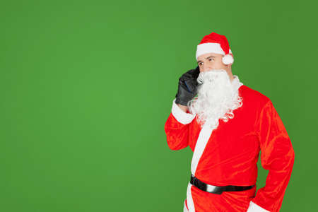 An adult Caucasian man dressed as Santa Claus is talking on his cell phone. The background is green.の写真素材