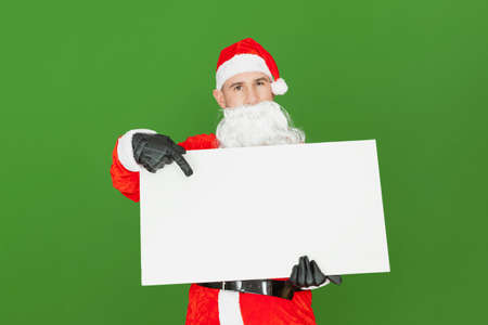 A Caucasian adult male dressed as Santa Claus is holding a blank white banner while pointing towards it with his index finger. The background is green.の写真素材