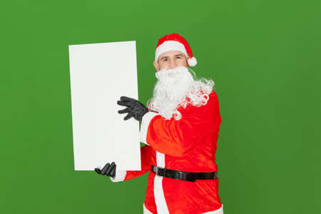An adult Caucasian male dressed as Santa Claus is holding a blank white panel vertically to the side of his body with his hands. The background is green.の写真素材