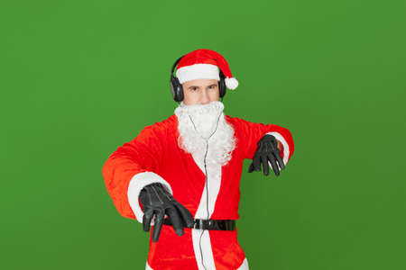An adult Caucasian man dressed as Santa Claus with headphones is imitating a disc jockey pretending to move the records and volume control in the air. The background is green.の写真素材