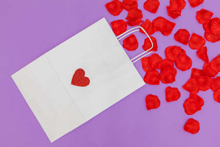 On a purple background is a white paper shopping bag on which is a red glitter heart. From the bag come out red petals, which are scattered.の写真素材