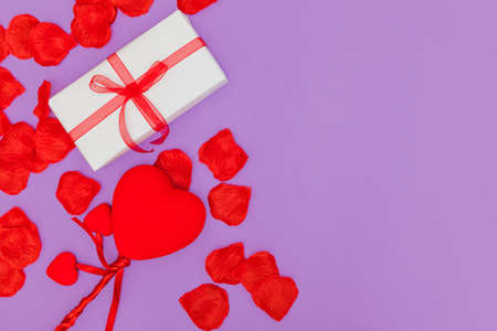 On a purple background there are several red petals, a gift wrapped with white paper and a ribbon with a bow and three hearts attached to a stick. On one side there is a lot of copy space.の写真素材
