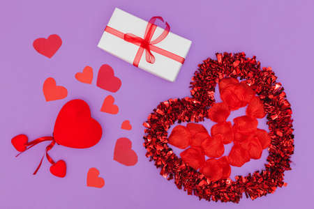 On a violet background there is a large bright heart, inside of which there are red petals. Next to it are smaller hearts and a gift box wrapped with white paper and a ribbon with a bow.の写真素材