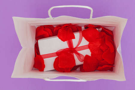 On a purple background is a white paper shopping bag and inside are red petals and a gift wrapped with white paper and a ribbon with a bow.の写真素材