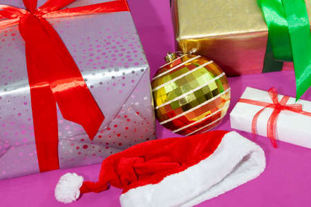 A close-up of a christmas items. It can see gifts with ribbon, a christmas golden ball, and a Santa hat, all over a pink background.の写真素材