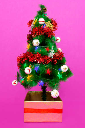 A small Christmas tree decorated with balls, stars, pine cones, garlands and colored lights. The base is inside a square box decorated with gold paper and a red ribbon. The background is pink.の写真素材