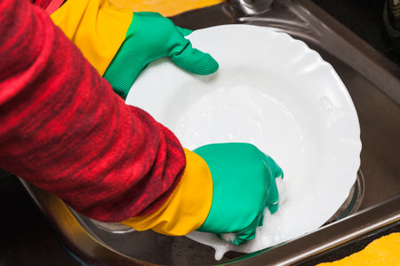 Close-up of the hands of a person wearing green and yellow rubber gloves scrubbing a white porcelain dish with a blue scouring pad in a sink using soap and water which generates foam.の写真素材