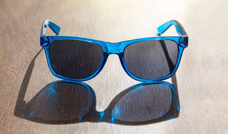 On a table there are sunglasses, with open temples, with blue frames. The light comes intense from...
