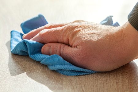 An older man's hand is wiping a wooden table with a blue cloth cloth. The light comes intense from the front and casts the shadow on the tabletop.の写真素材