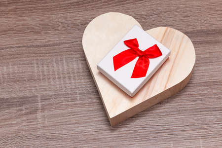 On a board is a wooden heart on top of which is a white gift box with a red ribbon.の写真素材