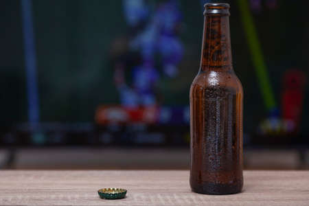 A bottle of fresh beer and a bottle cap on a wooden table. In the background, out of focus, there is a television with a sports broadcast.の写真素材