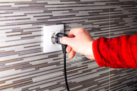 The hand of an unknown man plugging a black european plug into an electrical socket installed in a...