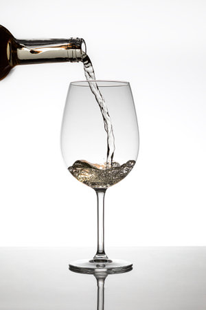 A bottle filling a glass of white wine. The liquid is in motion and remains frozen. The foot of the glass is reflected on the surface and the background is white.の写真素材