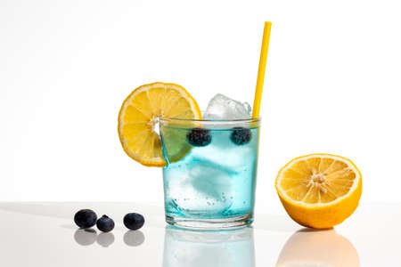 A glass of blue gin and tonic with ice, blueberries, a slice of lemon and a yellow straw. The background is white and the elements are reflected on the surface they are on.の写真素材