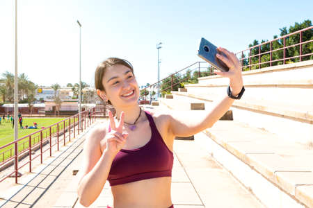 Smiling young Caucasian female athlete dressed in a purple top taking a selfie on a bleacher forming a V with her right hand. in the background is a sports area.の写真素材