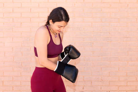 A young Caucasian woman dressed in leggings and a top putting on a pair of black boxing gloves. With one hand she is adjusting the glove of the other and in the background is a brick wall.の写真素材
