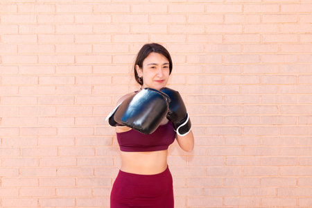 A young Caucasian woman wearing black boxing gloves is punching in front. The girl's face is in focus. In the background is a brick wall.の写真素材
