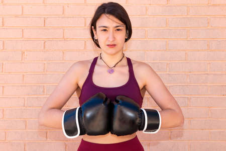 A serious young Caucasian woman with boxing gloves is putting her fists together in front of her chest. The background is a brick wall.の写真素材