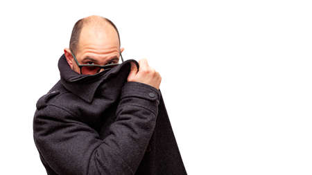 A bald adult male wearing sunglasses hides behind his dark jacket and peers over his glasses. Behind the man the background is white.の写真素材