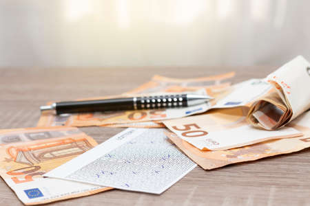 On a wooden table there is a lottery ticket, a pen and several 50-euro bills scattered and rolled up...