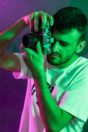 Portrait of a young bearded Caucasian man posed taking pictures with an antique camera framed vertically. It is illuminated with green and magenta colored lights.の写真素材