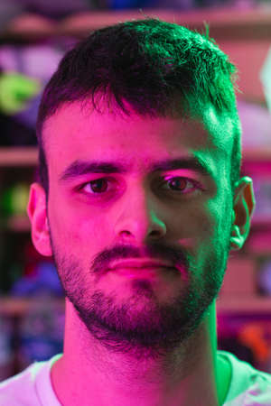 Portrait of a young Caucasian man with beard illuminated with green and magenta colored lights. In...
