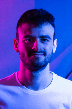 Portrait of a young Caucasian man with beard dressed in a white T-shirt illuminated with blue and...