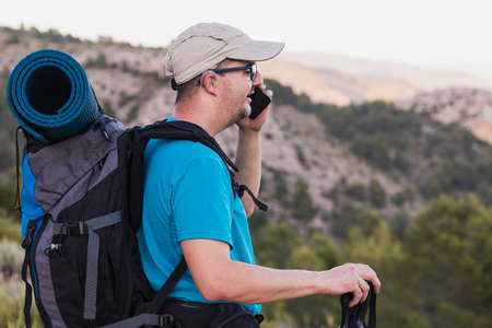 A hiker, adult, male, Caucasian, with a backpack, mat, poles, dressed in a blue T-shirt, cap and gray pants wearing a hearing aid and glasses is talking on the mountain on his cell phone.の写真素材