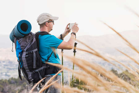 A hiker, adult, male, Caucasian, with a backpack, mat, poles, dressed in a blue T-shirt, cap and gray pants wearing a hearing aid and glasses is on the mountain taking a picture with his cell phone.の写真素材