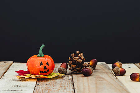 Halloween pumpkin on leaves that are on top of some wooden boards where there is also a pine cone and acorns. The background is dark.の写真素材