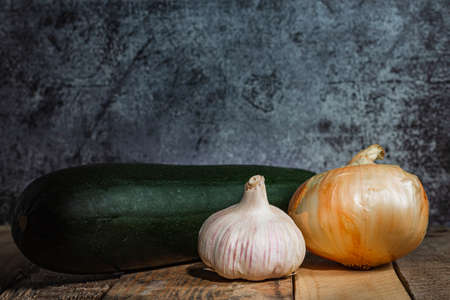 Still life composed of a head of garlic, a sweet onion and a large zucchini placed on a table with old wooden boards and an abstract background of a gray wall in a dark environment.の写真素材