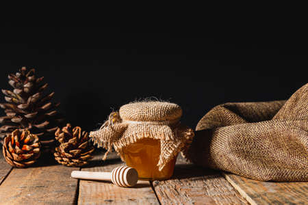 Rustic still life in which you can see a glass jar with honey covered with a sackcloth, next to it a wooden honey stick and some pine cones. Everything is on a table of old wooden planks.の写真素材