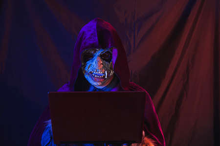 Portrait of a zombie dressed in shirt and hooded cape using his laptop. The scene is dark, lit by...