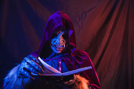 Portrait of a zombie dressed in a shirt and hooded cloak turning the pages of a diary or journal while looking at the book. The scene is dark, illuminated by blue and orange lights.の写真素材